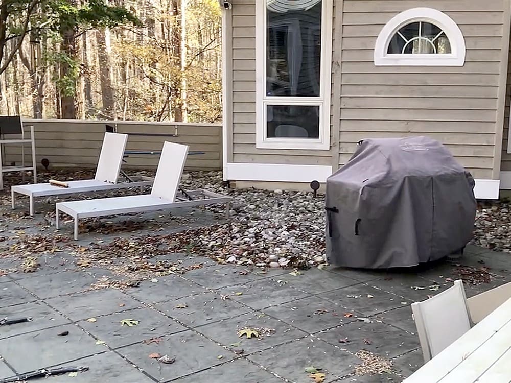 A patio with tile flooring featuring two modern white lounge chairs on the left. A covered grill is positioned to the right of the lounge chairs. The background shows a gray wooden house with large windows and a circular decorative window above. Trees and autumn leaves are scattered around.